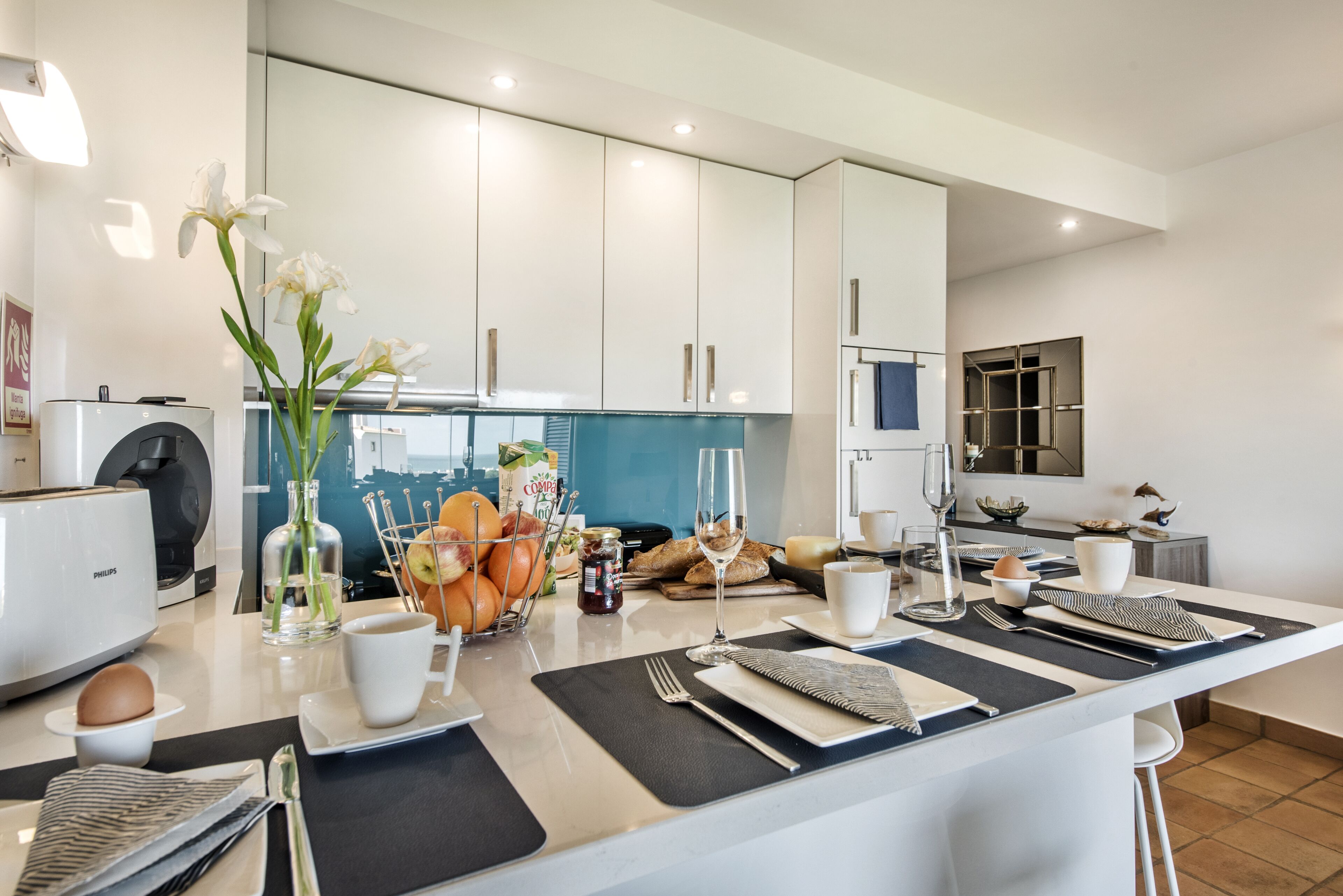 A modern kitchen with white cabinets, a blue backsplash, and a breakfast setup including fruits, bread, and a coffee maker.