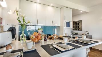 A modern kitchen with white cabinets, a blue backsplash, and a breakfast setup including fruits, bread, and a coffee maker.