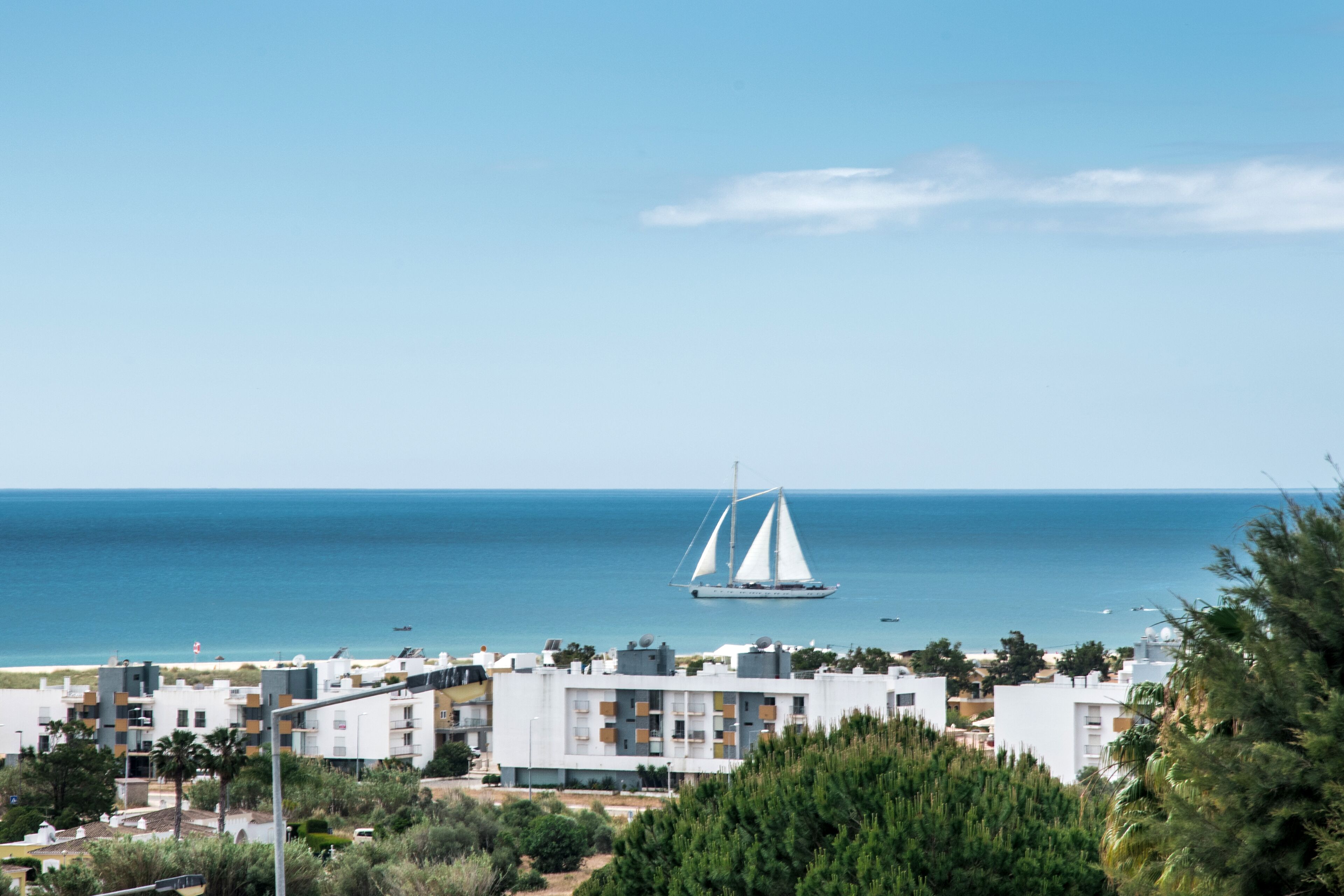 A coastal town with modern buildings, a sailboat on the water, and a clear view of the sea.