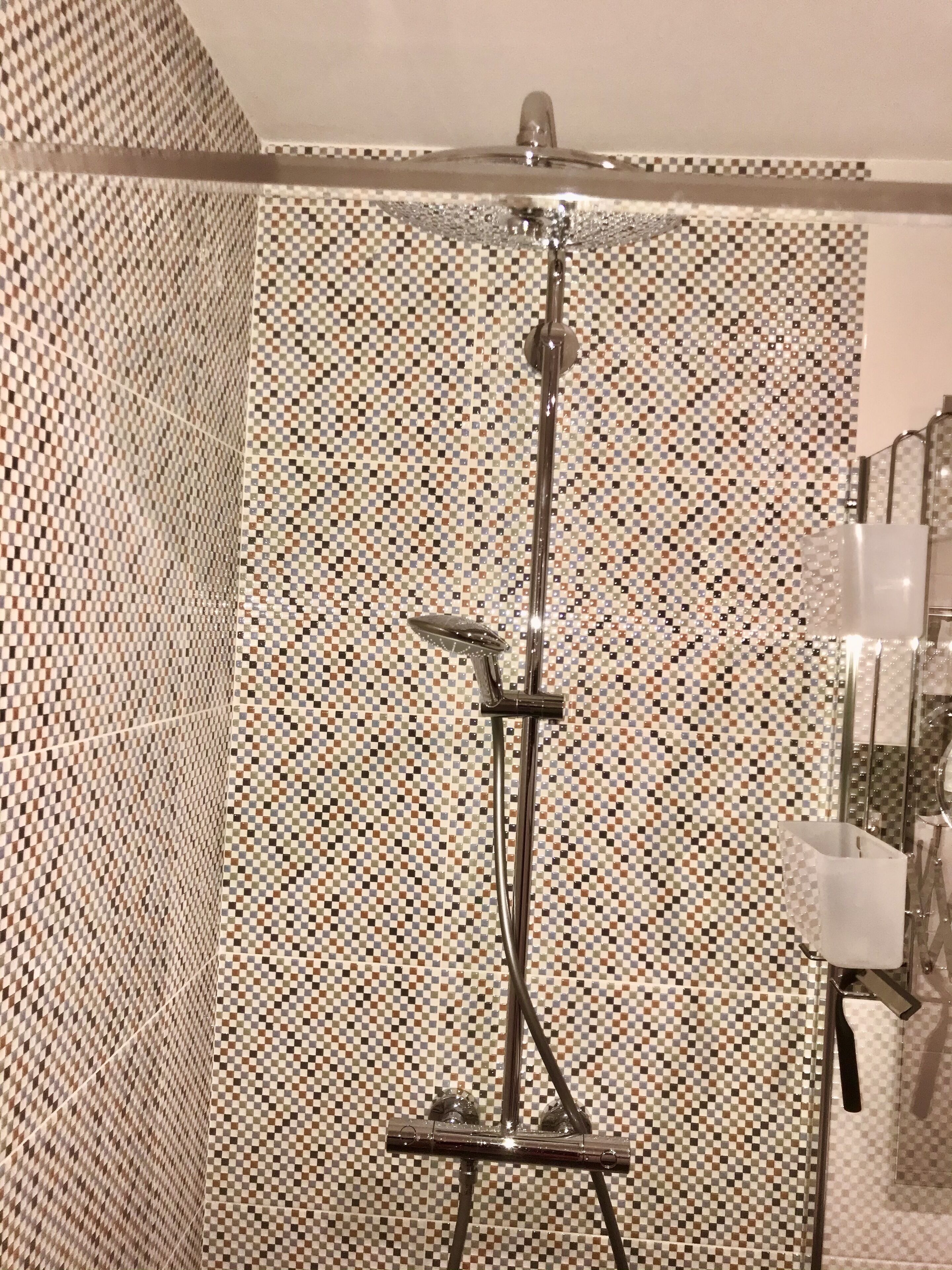 A shower with a checkered tile wall, a handheld showerhead, and a handheld shower attachment.