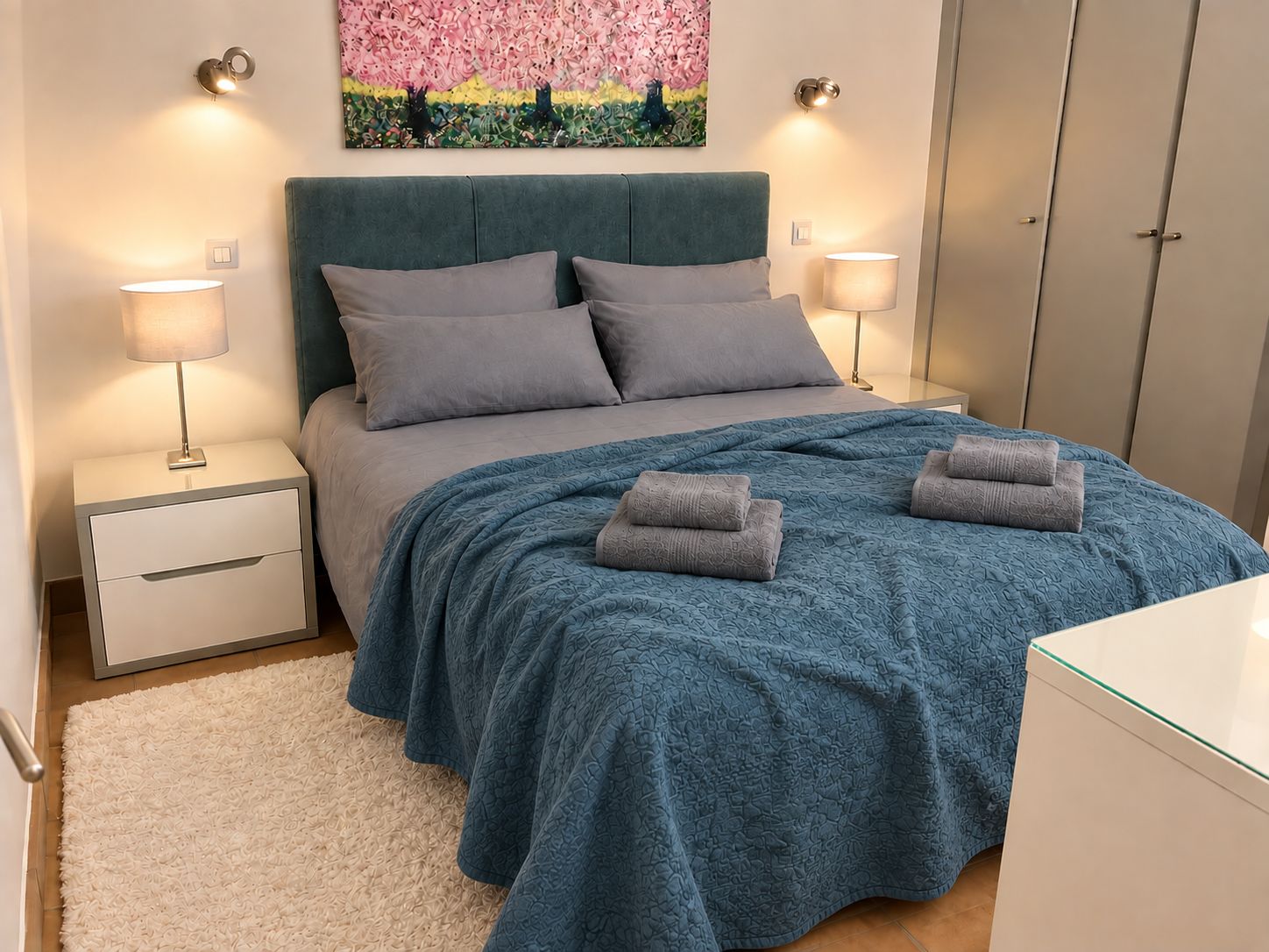Un dormitorio con cama, dos lámparas de noche, una mesita, un cuadro en la pared y un armario.