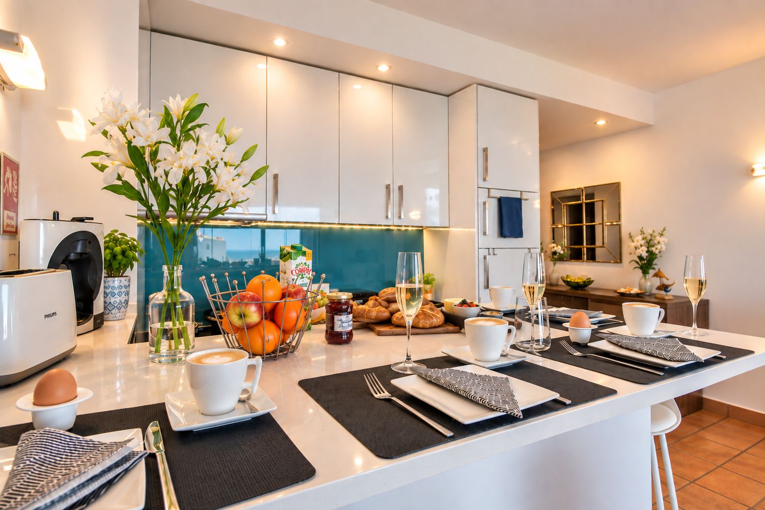 A modern kitchen with a central island, a coffee machine, and a breakfast setting with croissants, fruit, and wine glasses.