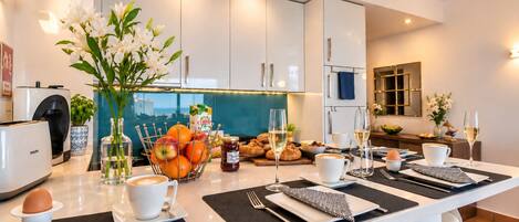 A modern kitchen with a central island, a coffee machine, and a breakfast setting with croissants, fruit, and wine glasses.