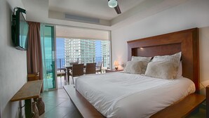 A hotel room with a large bed, a ceiling fan, a TV mounted on the wall, and a balcony with a view of a high-rise building. - Wake Up To Sounds Of The Ocean Condo (Puerto Vallarta)