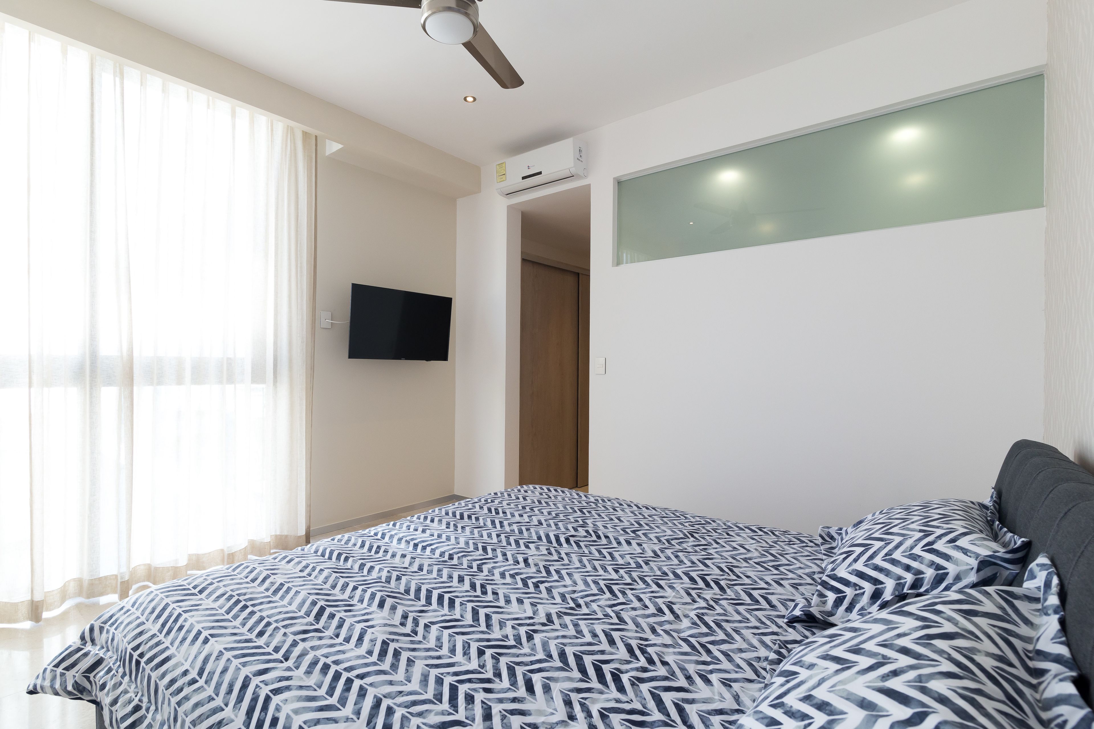 Quarto com cama, ventoinha no teto, ar condicionado, televisão e janela com cortinas.