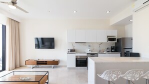 Una cocina moderna con armarios blancos, un microondas integrado y una cafetera. Hay un comedor con una mesa de vidrio y sillas blancas.