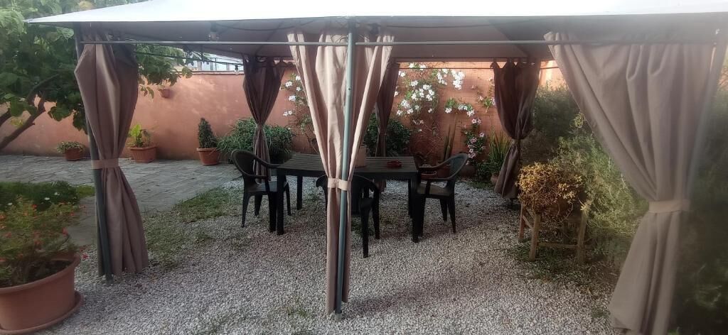 A covered outdoor seating area with a table and chairs, surrounded by potted plants and a gravel ground.