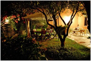 Un espace extérieur aménagé pour les repas, avec une table et des chaises couvertes, entouré de verdure et d’un arbre.