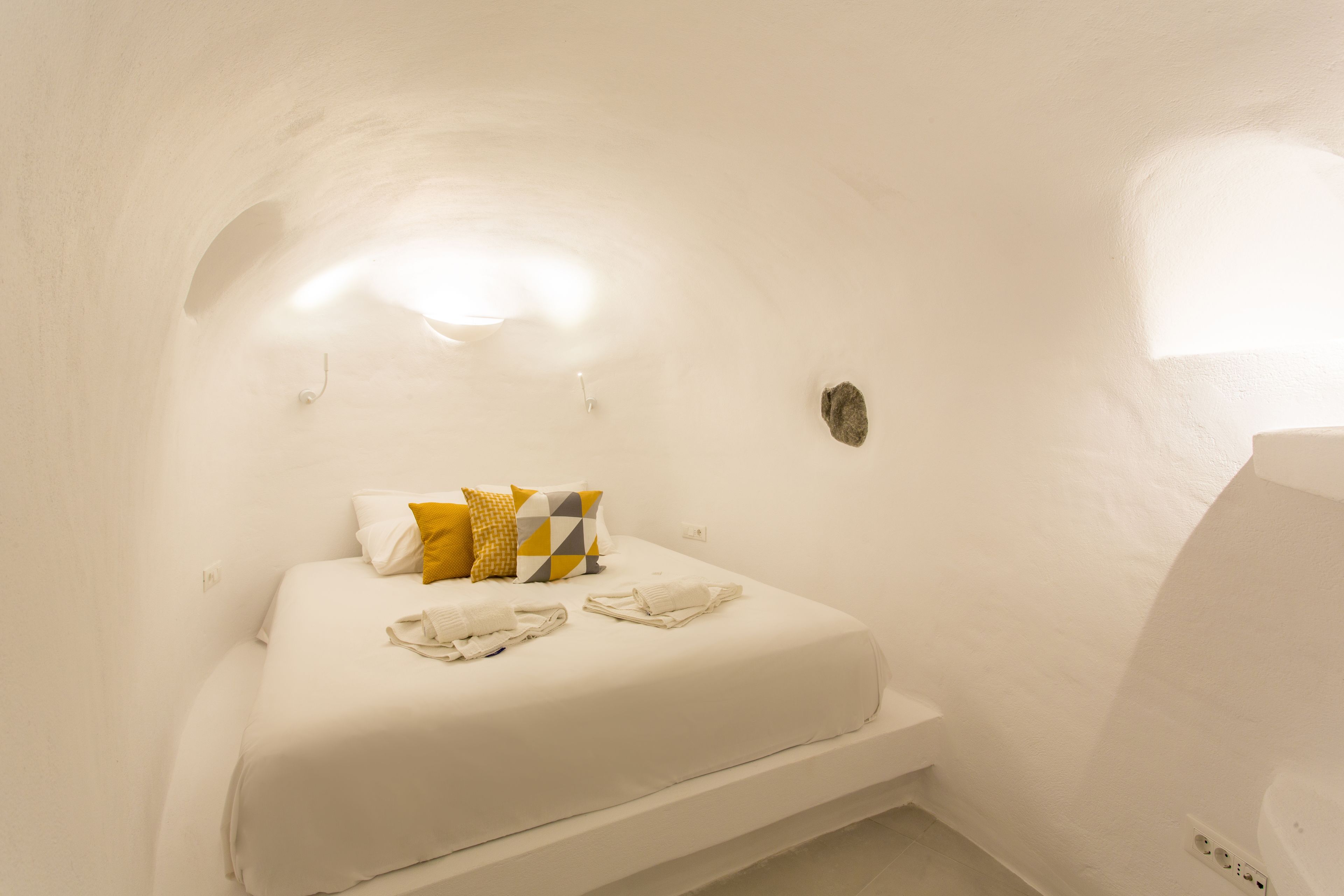 Una cama individual con colchón blanco, almohada de diseño amarillo y gris, y dos toallas dobladas. La habitación tiene paredes blancas y un techo inclinado.