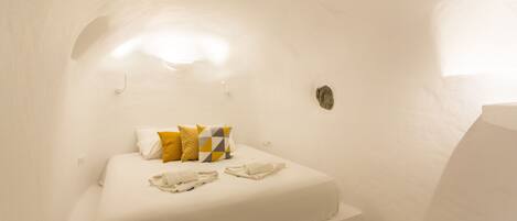 Una cama individual con colchón blanco, almohada de diseño amarillo y gris, y dos toallas dobladas. La habitación tiene paredes blancas y un techo inclinado.