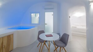 Una habitación moderna y compacta, con una paleta de colores blanco, que incluye una mesa redonda con dos sillas, una ventana pequeña y una bañera empotrada.