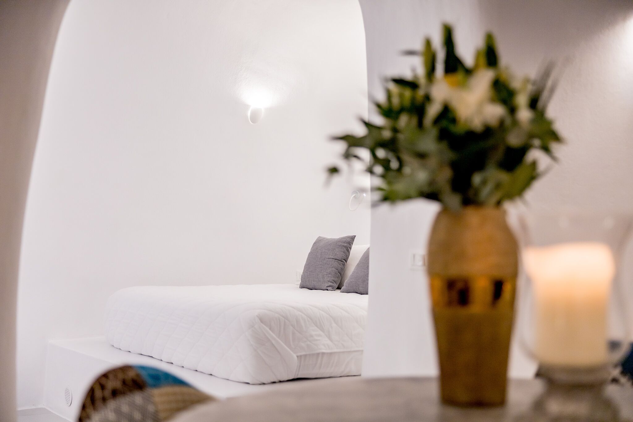 A white, minimalist bedroom with a bed, a vase with flowers, and a lit candle.
