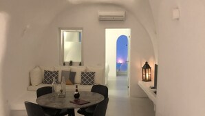 Una habitación moderna con paredes blancas, una mesa redonda de comedor y sillas, un sofá con cojines estampados, y vistas al atardecer a través de un arco.
