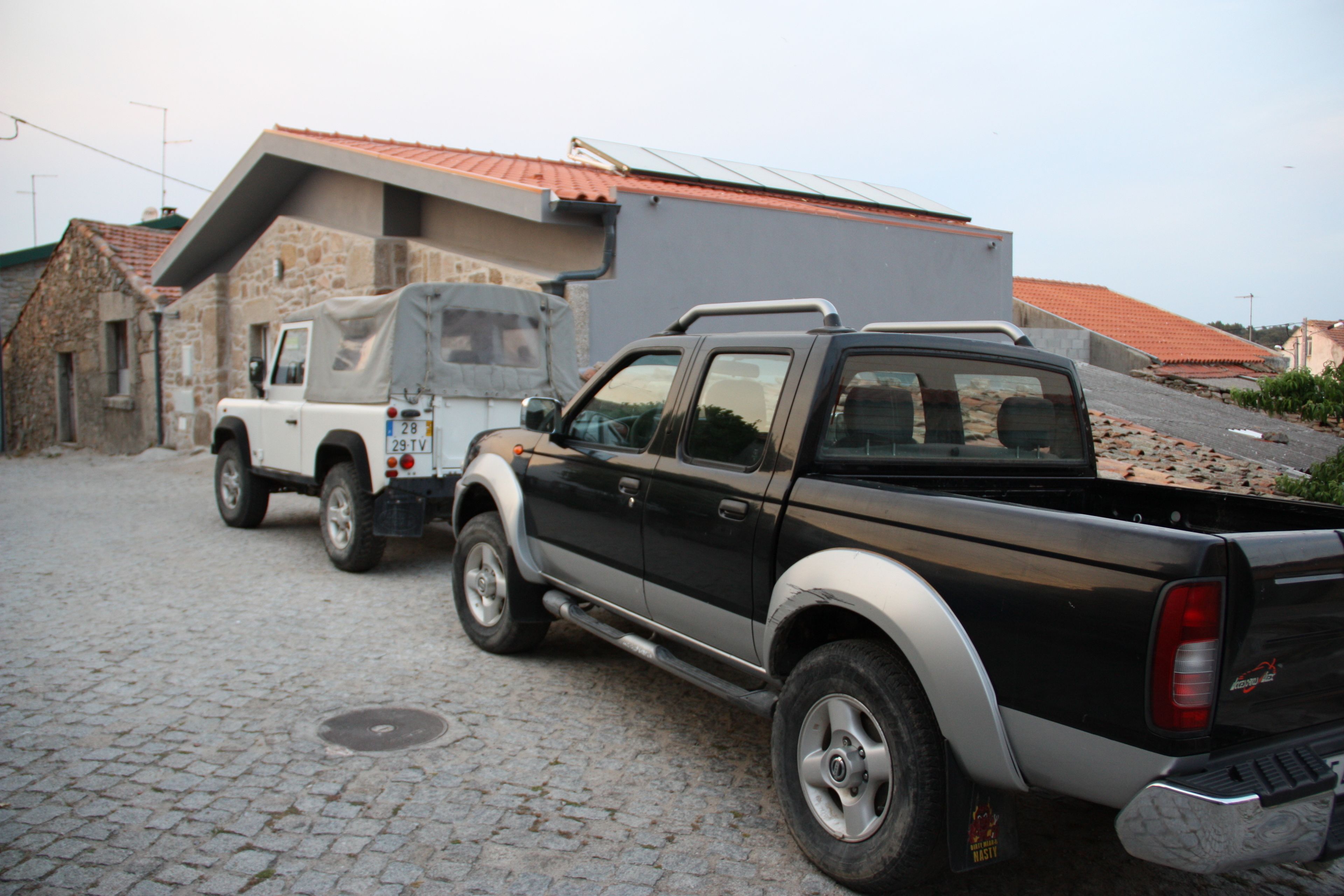 Una camioneta negra estacionada en una calle adoquinada junto a una camioneta blanca y un edificio con tejado de tejas rojas.