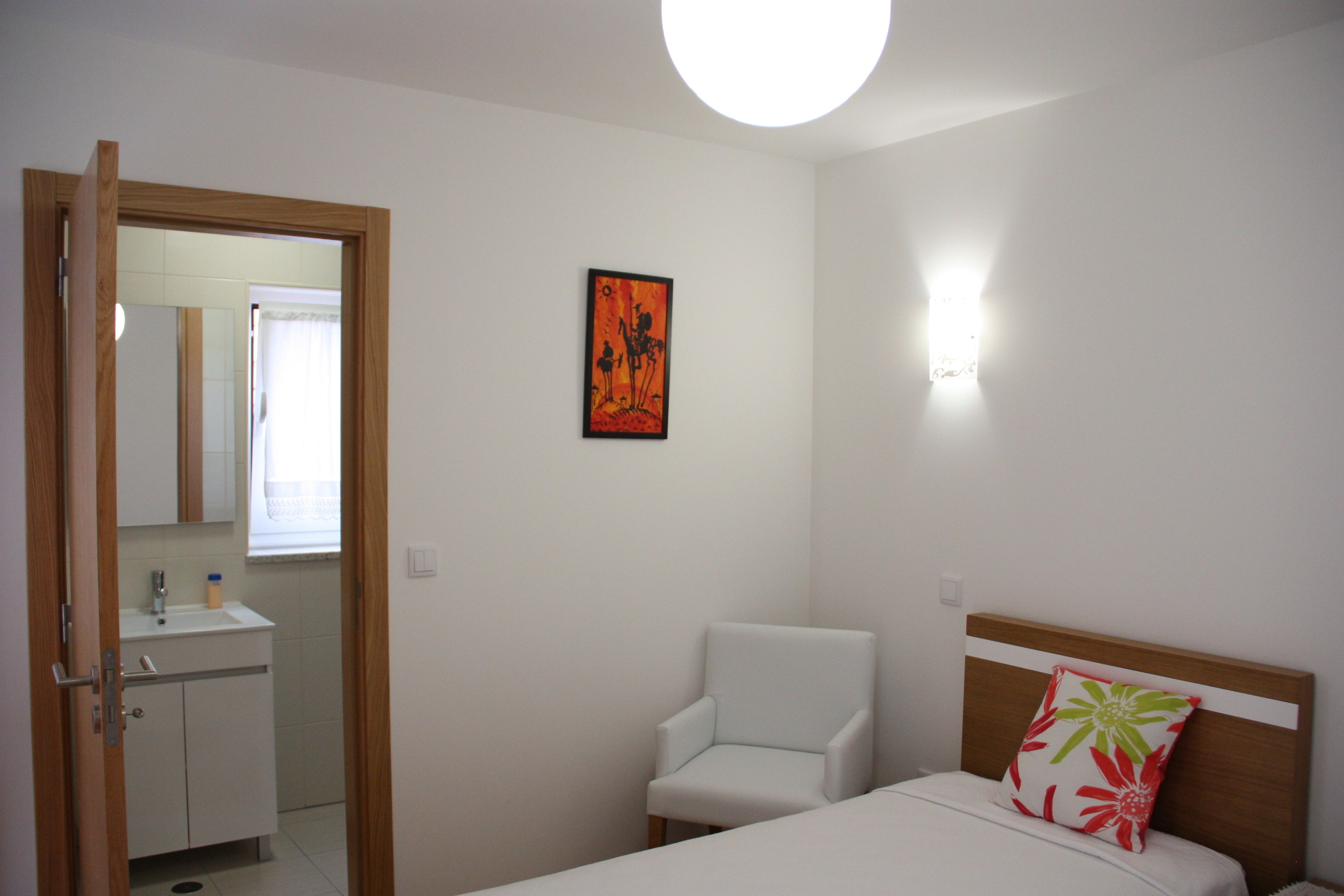 Un dormitorio con cama, una silla, una mesita de noche, una lámpara de pared y un cuadro.