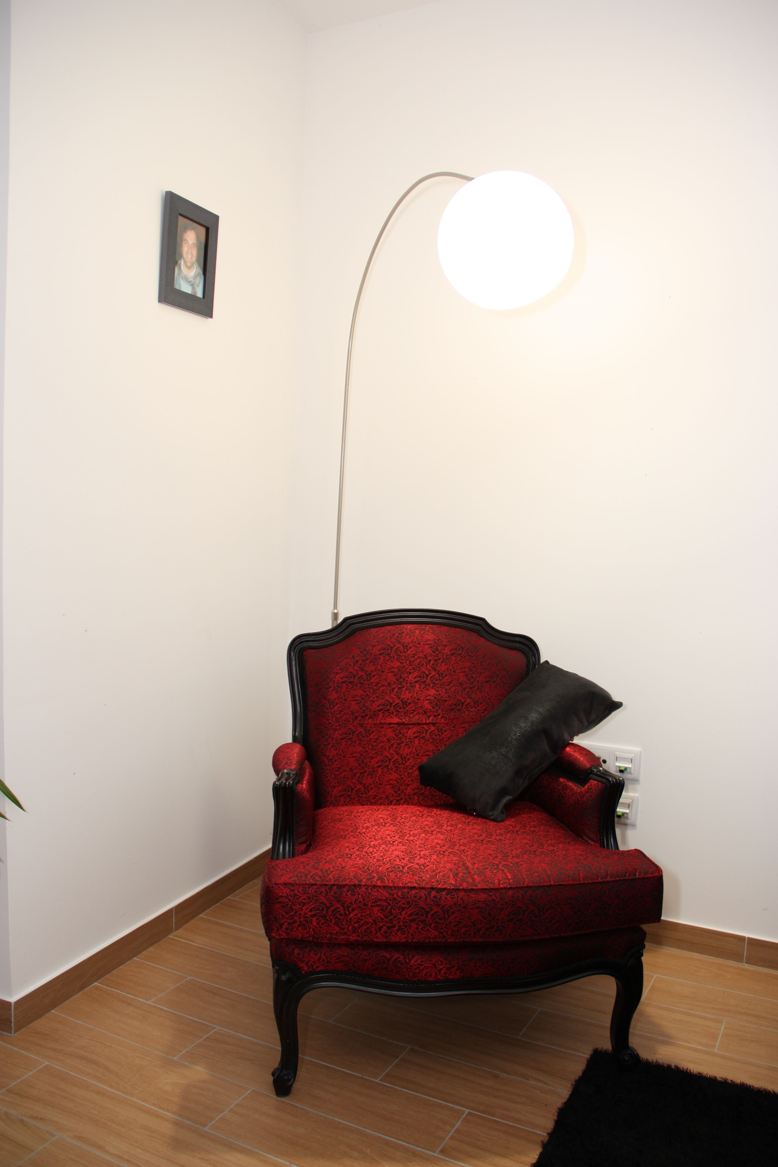 A red upholstered chair with a black cushion, a floor lamp, and a small framed picture on the wall.