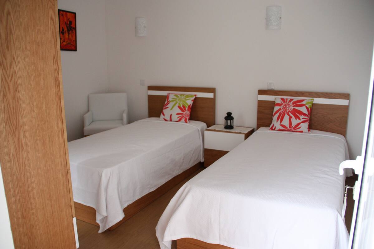 A room with two single beds, a chair, a nightstand, and a wall-mounted lamp.