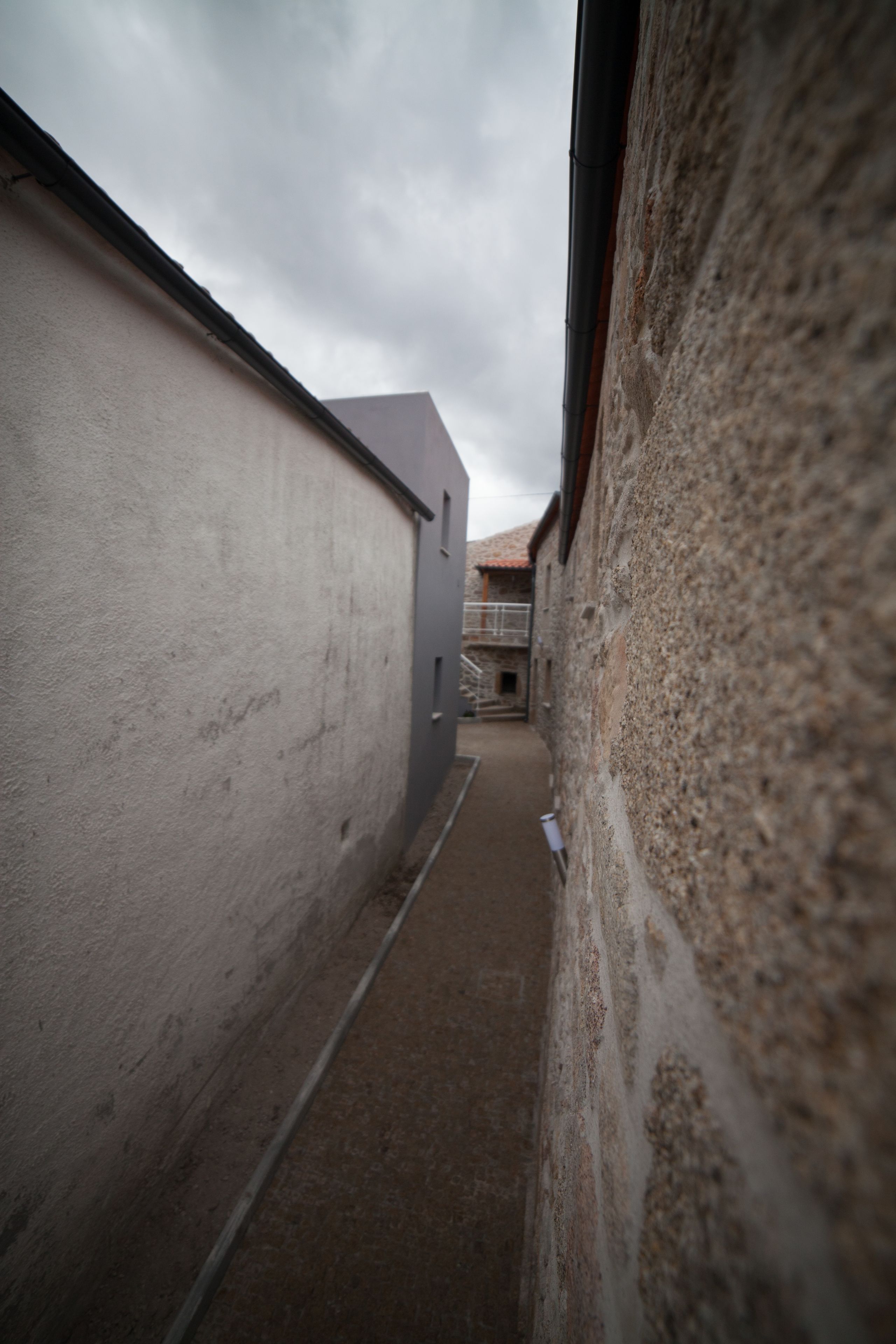 A narrow alleyway between buildings with a rough-textured wall on the right and a smooth-textured wall on the left.