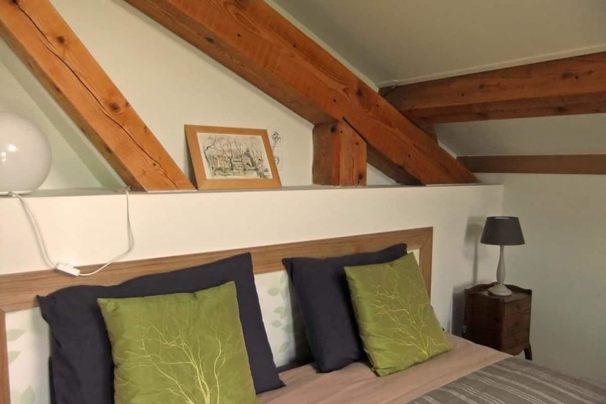 Ein Schlafzimmer mit Holzbalkendecke, einem Bett mit Kopfteil, einer Nachttischlampe und einem gerahmten Bild an der Wand.