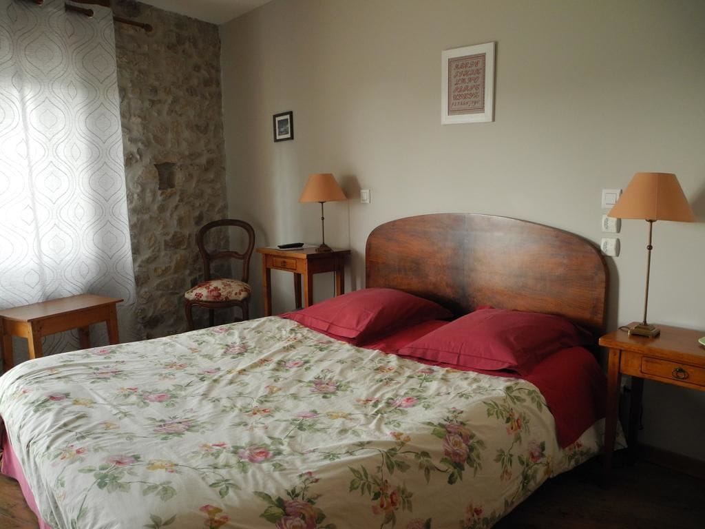 A bedroom with a wooden bed, red pillows, a floral bedspread, a stone wall, a chair, two bedside tables with lamps, and a framed picture on the wall.