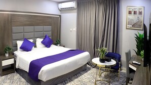Habitación de hotel con una cama grande, una mesita redonda pequeña, una silla azul y una planta en maceta.