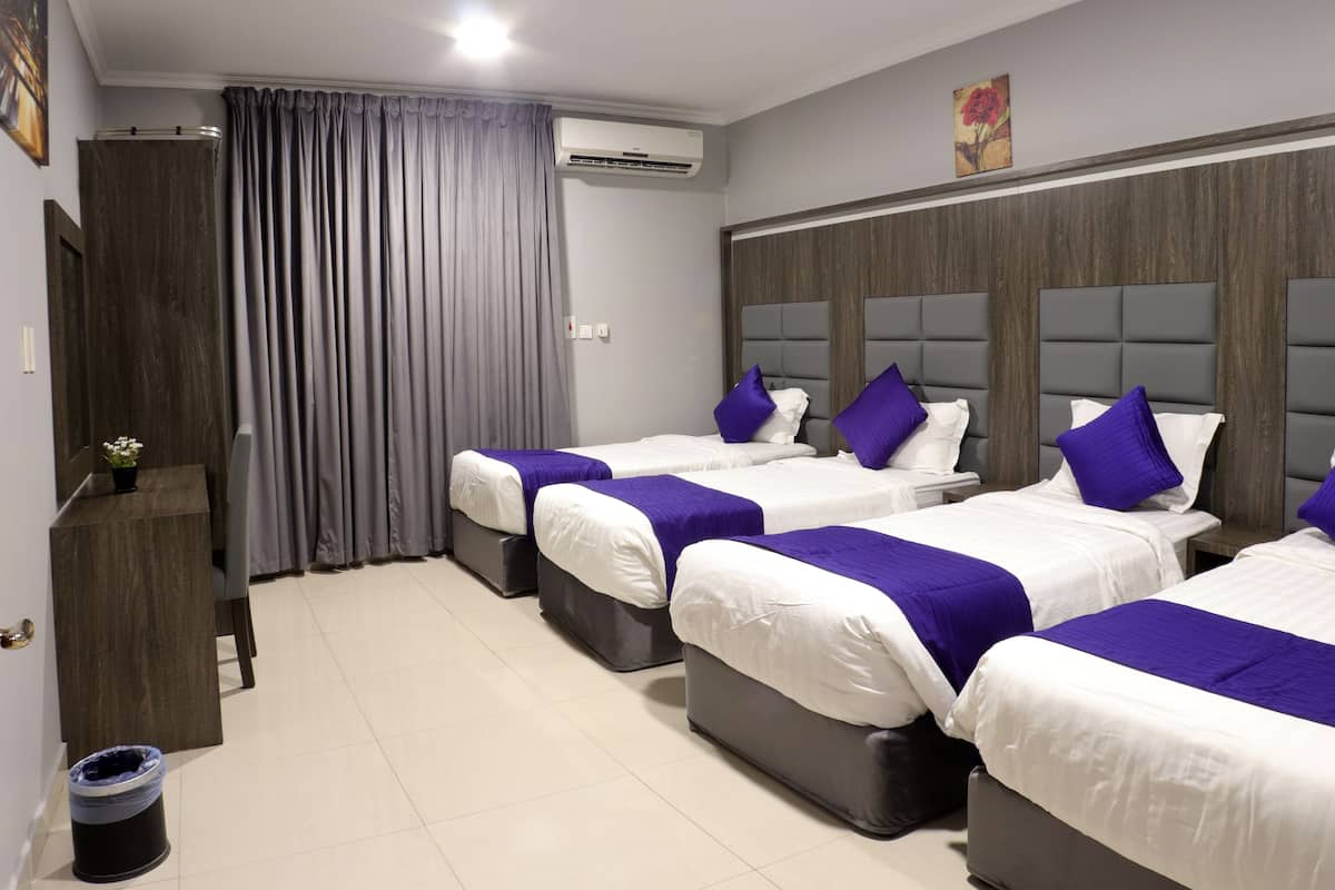 Una habitación de hotel con cuatro camas, cada una con ropa de cama blanca y detalles morados. Tiene un cabecero de madera, una cortina gris y un cubo de basura pequeño.