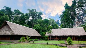 Two thatched-roof huts with wooden walls, surrounded by lush greenery and trees.