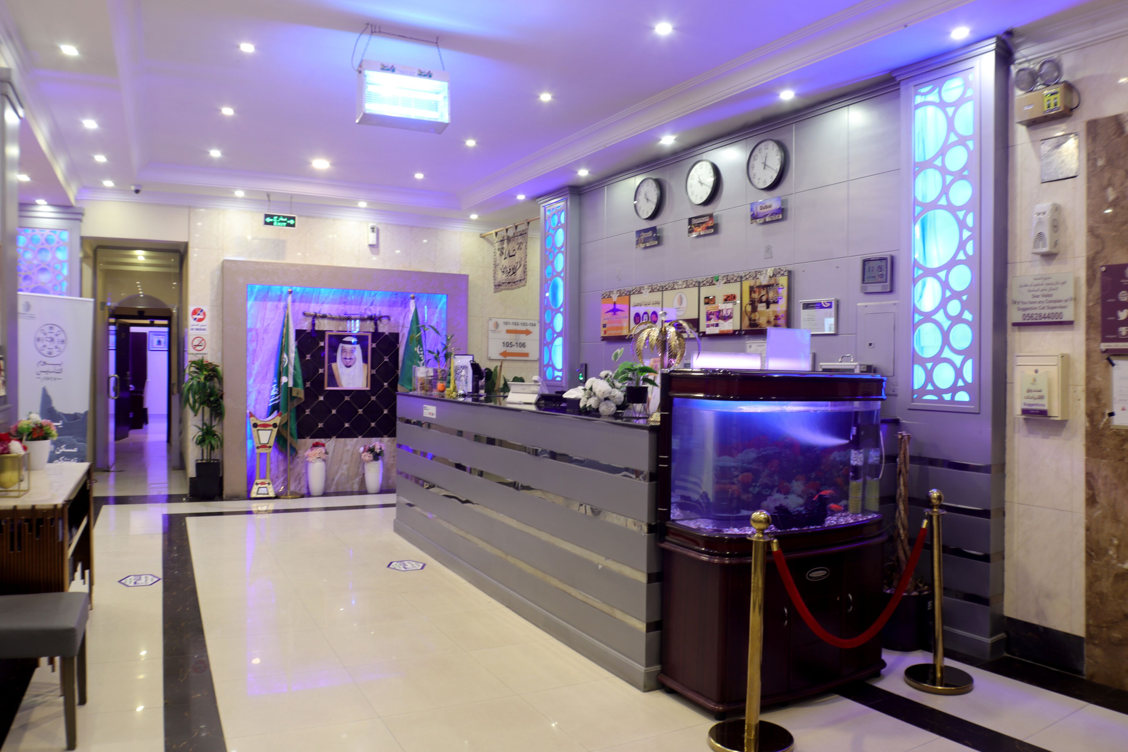 A hotel reception area with a reception desk, a fish tank, clocks on the wall, and a plant.