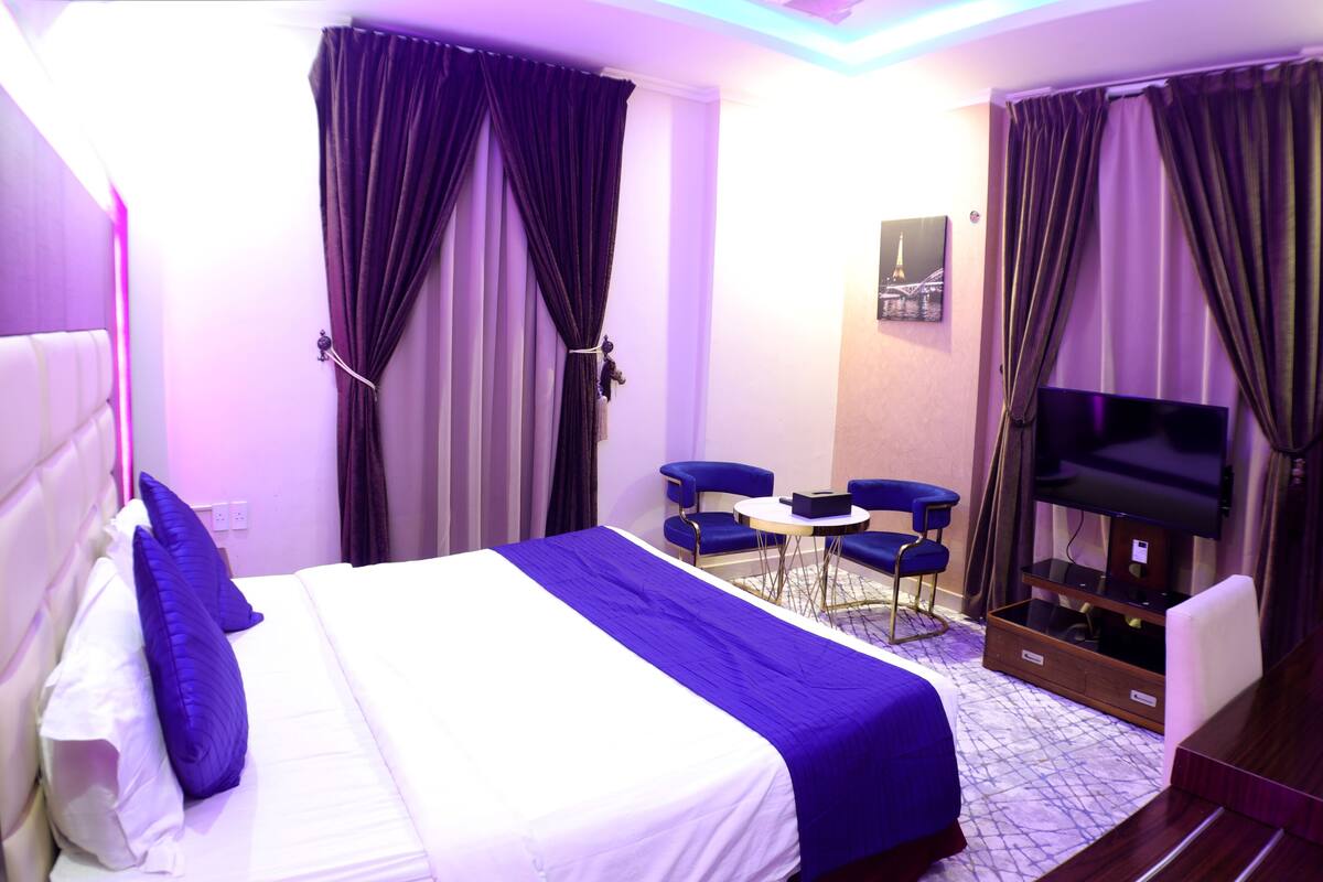 Una habitación de hotel con una cama, dos sillas azules, una mesita, un televisor y cortinas.