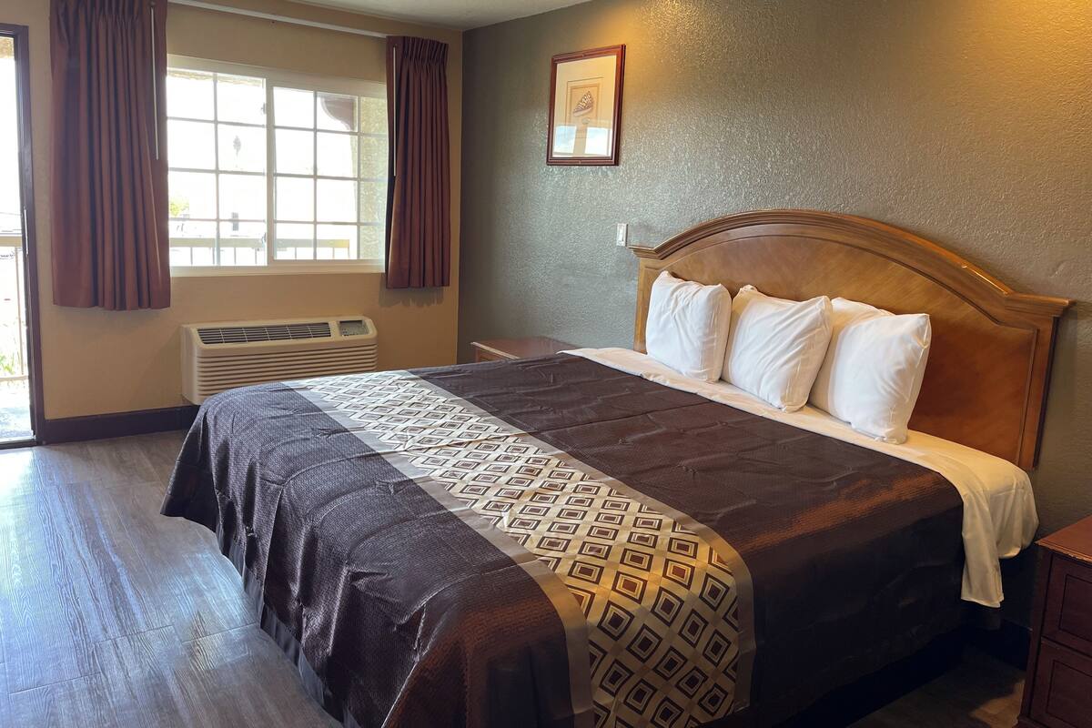 A hotel room with a large bed, a window with curtains, a wall-mounted picture, and a wooden nightstand.