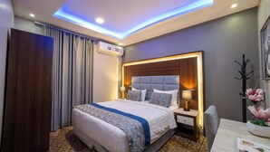 Quarto de hotel moderno com uma cama grande, mesa de cabeceira com abajur, cabeceira com iluminação embutida e iluminação no teto embutida.