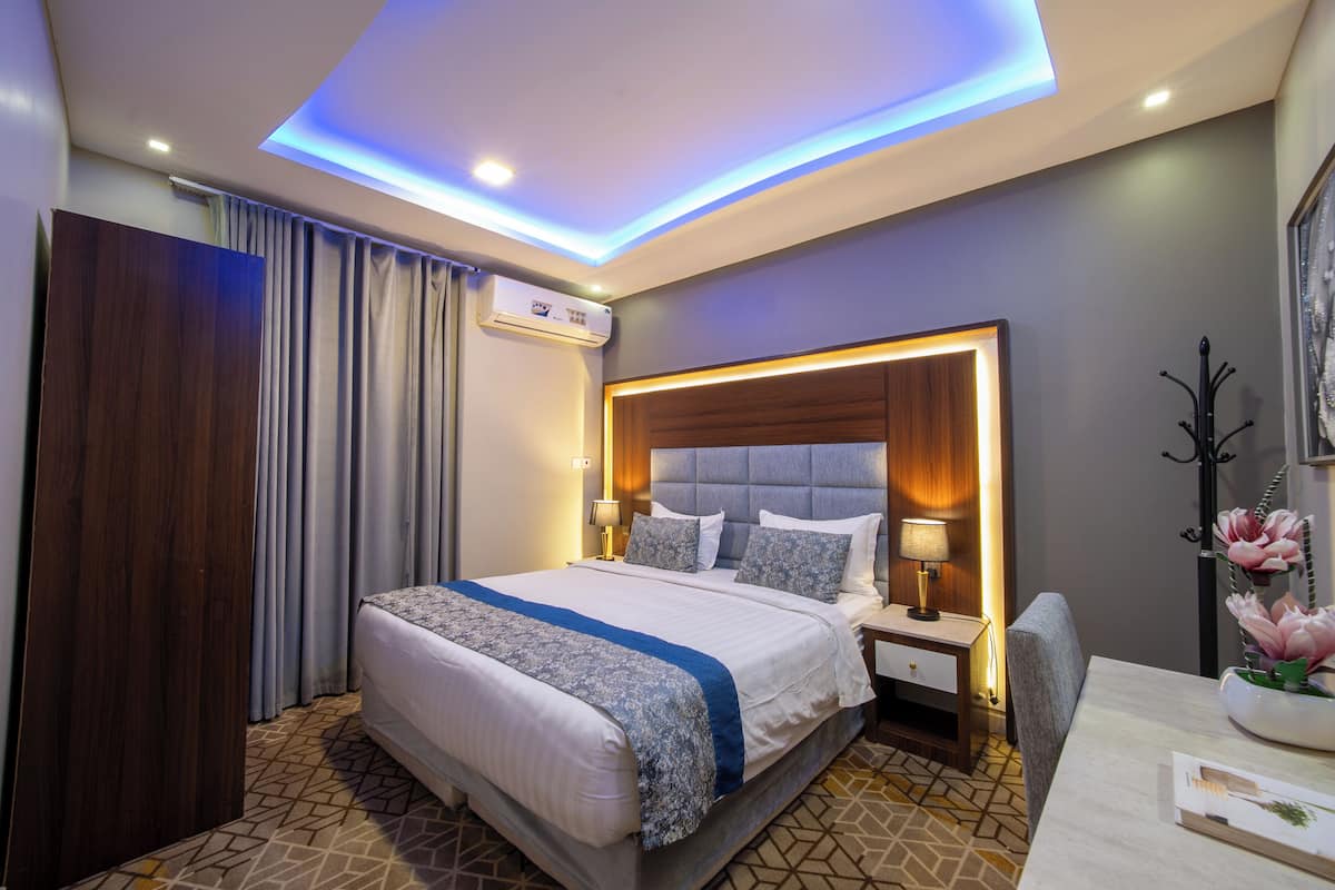 A modern hotel room with a large bed, a bedside table with a lamp, a headboard with built-in lighting, and a ceiling with recessed lighting.