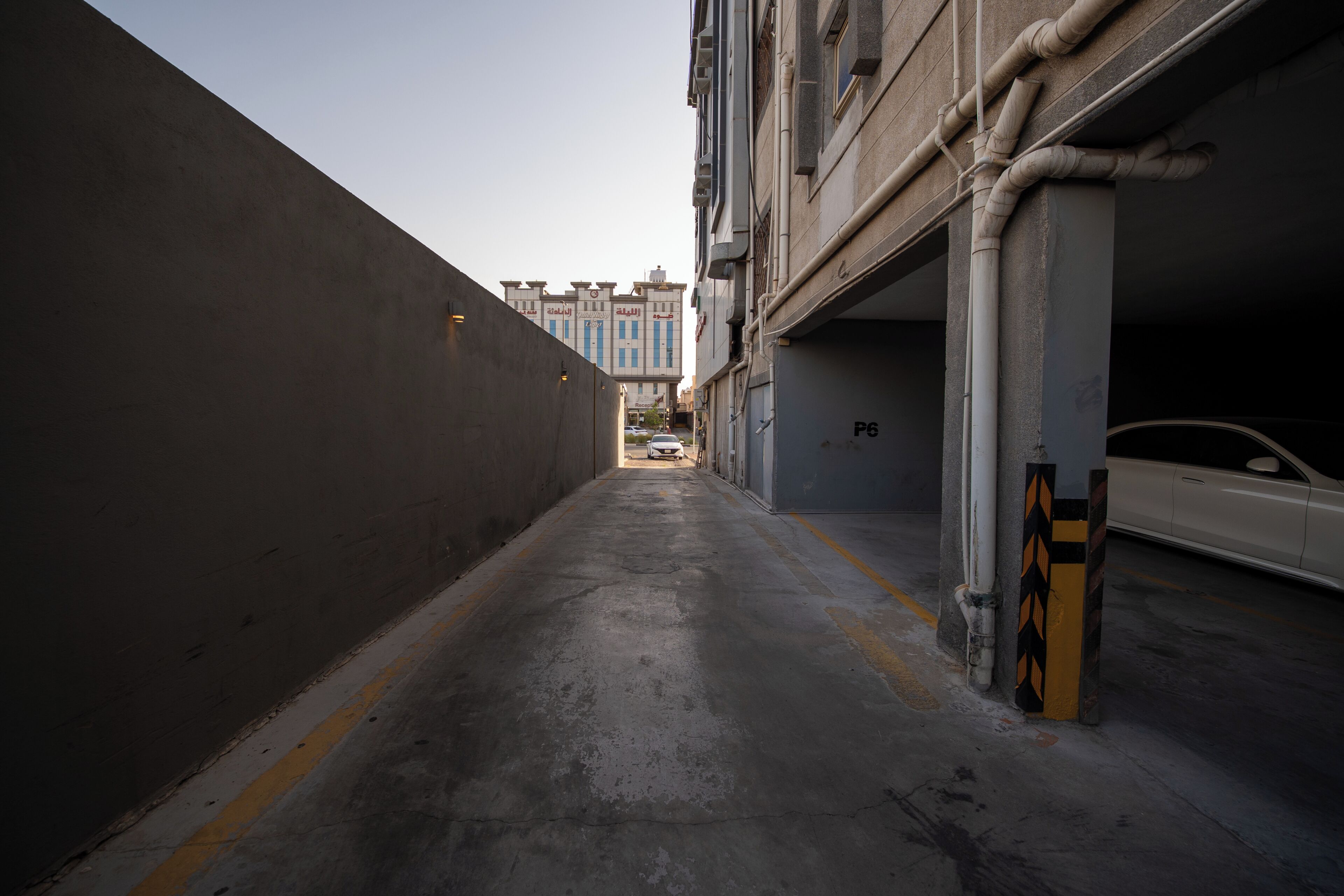 Ein schmaler Stadtgang mit Betonboden, ein weißes Auto parkt rechts und ein Gebäude mit blauen Fenstern im Hintergrund.