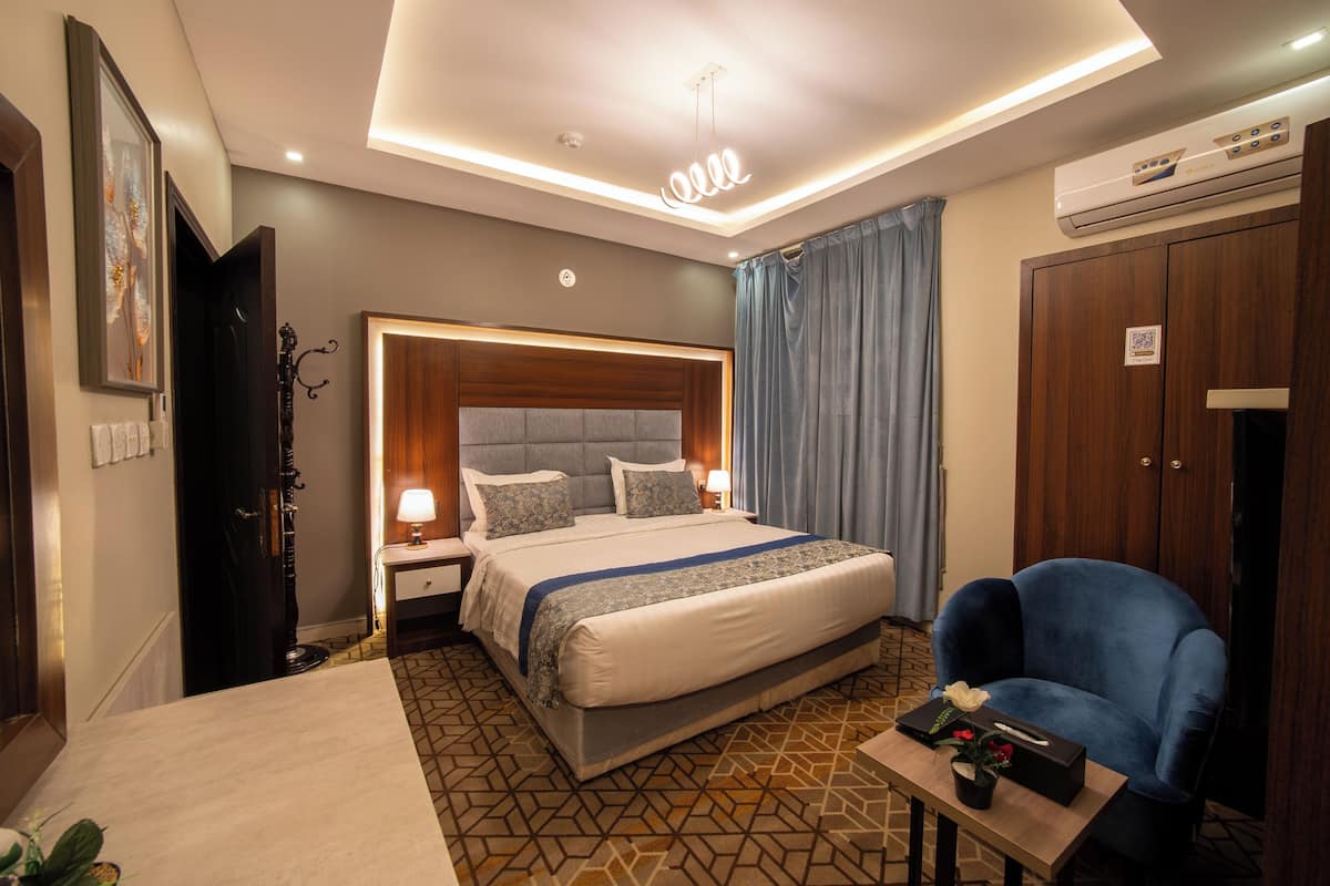 A hotel room with a large bed, a bedside table, a chair, a small table with a vase, and an air conditioning unit.