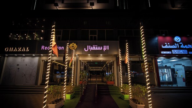 Entrada de hotel com balcão de recepção, plantas em vasos e pilares iluminados.