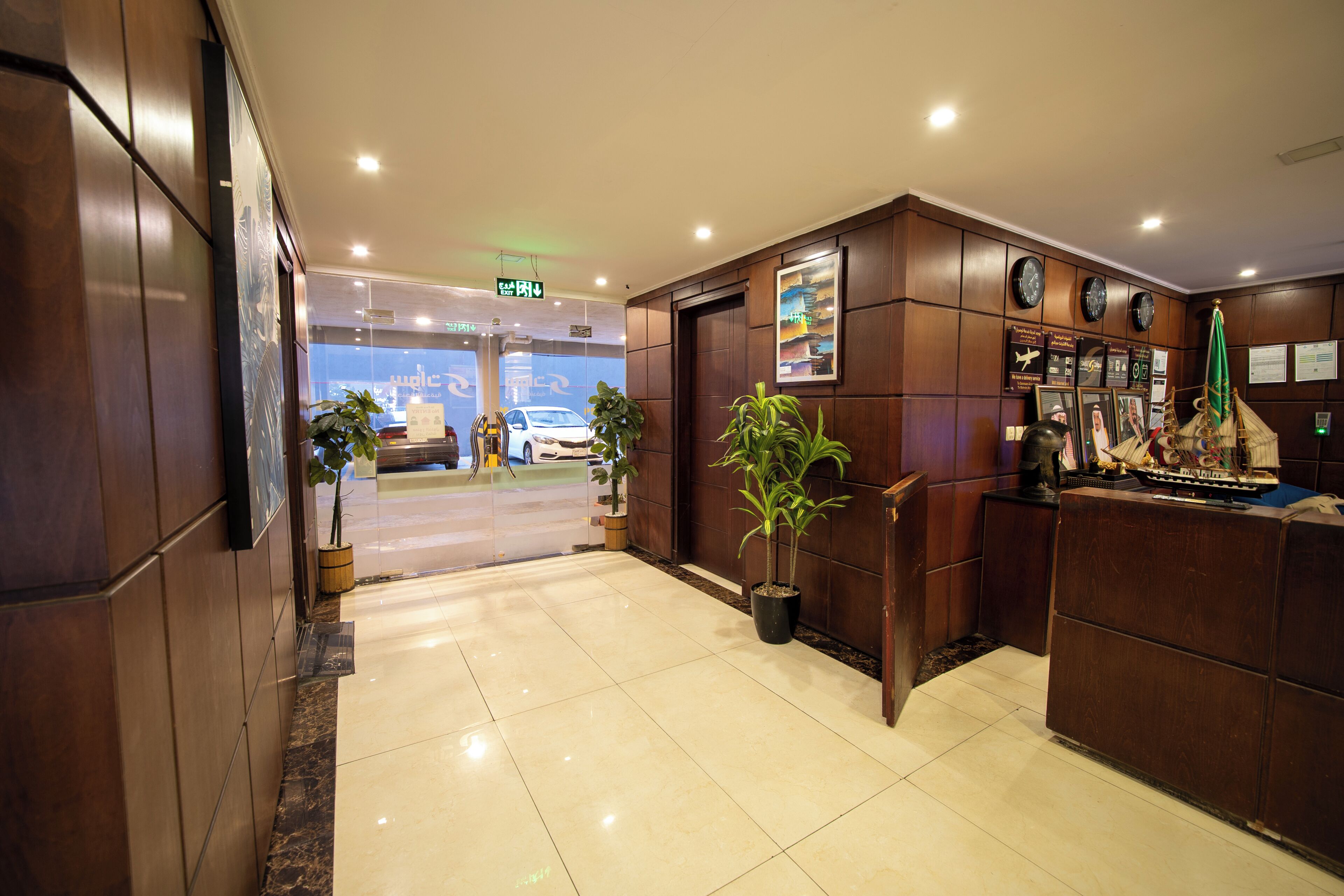 A hotel lobby with a reception desk, potted plants, clocks on the wall, and a car visible through the glass door.