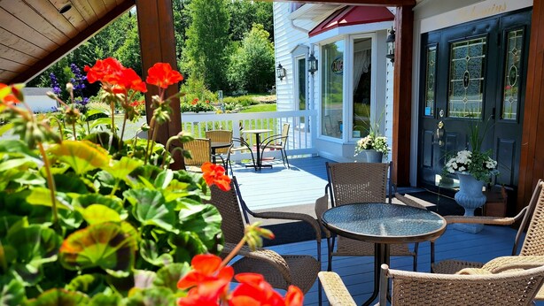 A covered patio with wicker furniture, a glass door entrance, and a view of a lush garden. - Auberge Les Etchemins (Lac-Etchemin)
