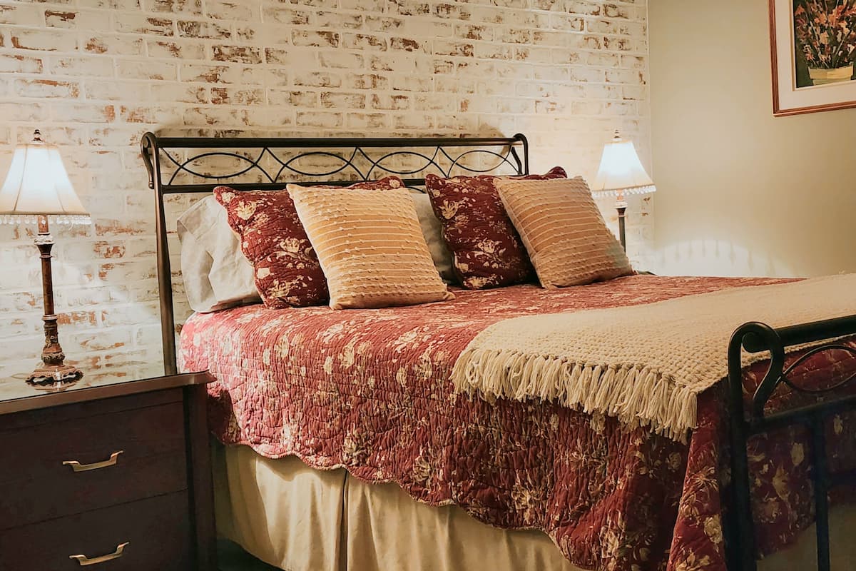 Une chambre avec un mur décoré d’un motif en briques, un cadre de lit en métal, une couvre-lit rouge et beige, deux lampes de chevet et une table de chevet avec des tiroirs.