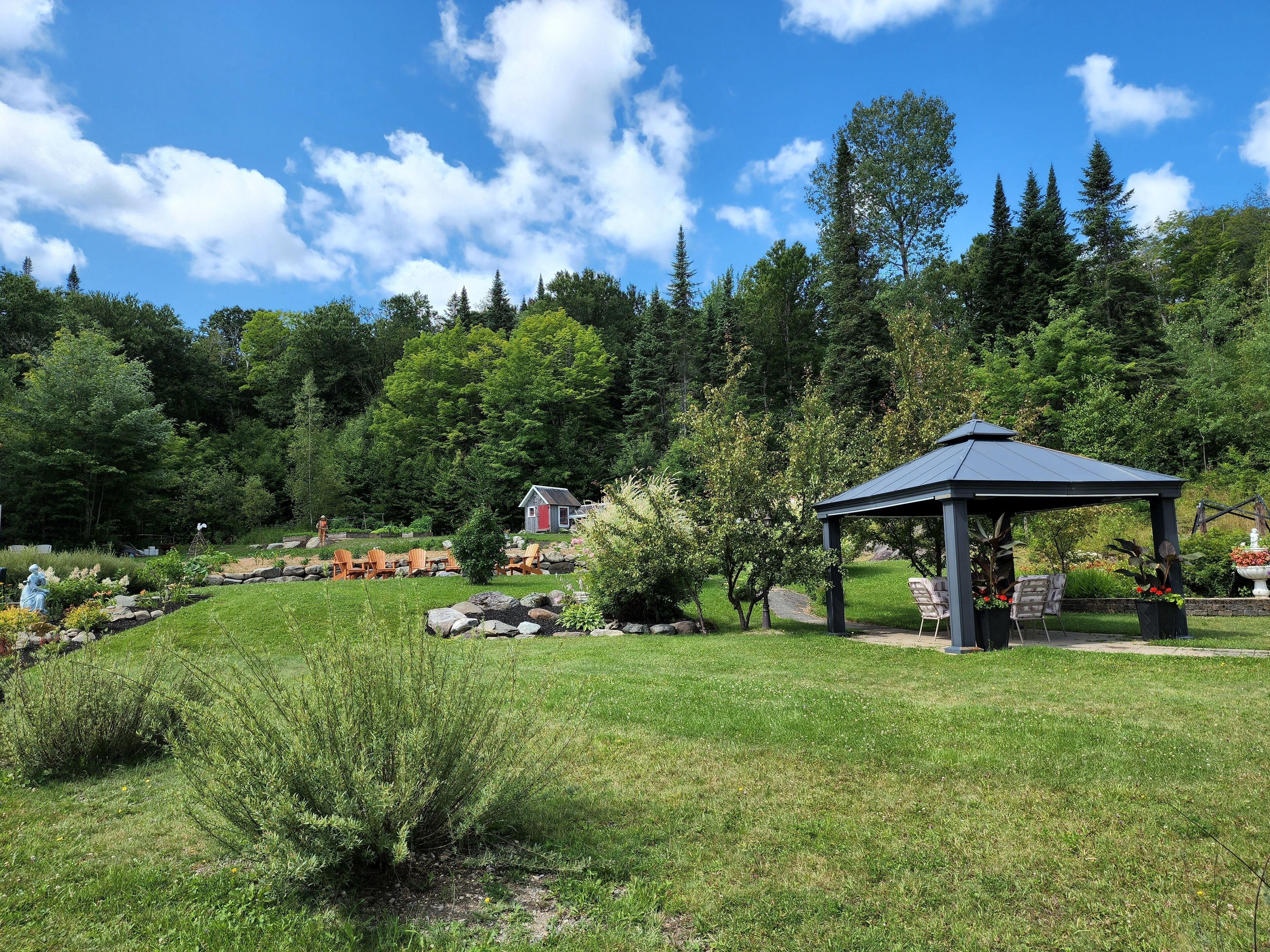 A gazebo in a lush garden with a pond, surrounded by trees and a clear sky.