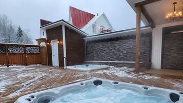 Uma banheira de hidromassagem em um quintal nevado com um deck de madeira, uma parede de pedra e uma casa com telhado vermelho.