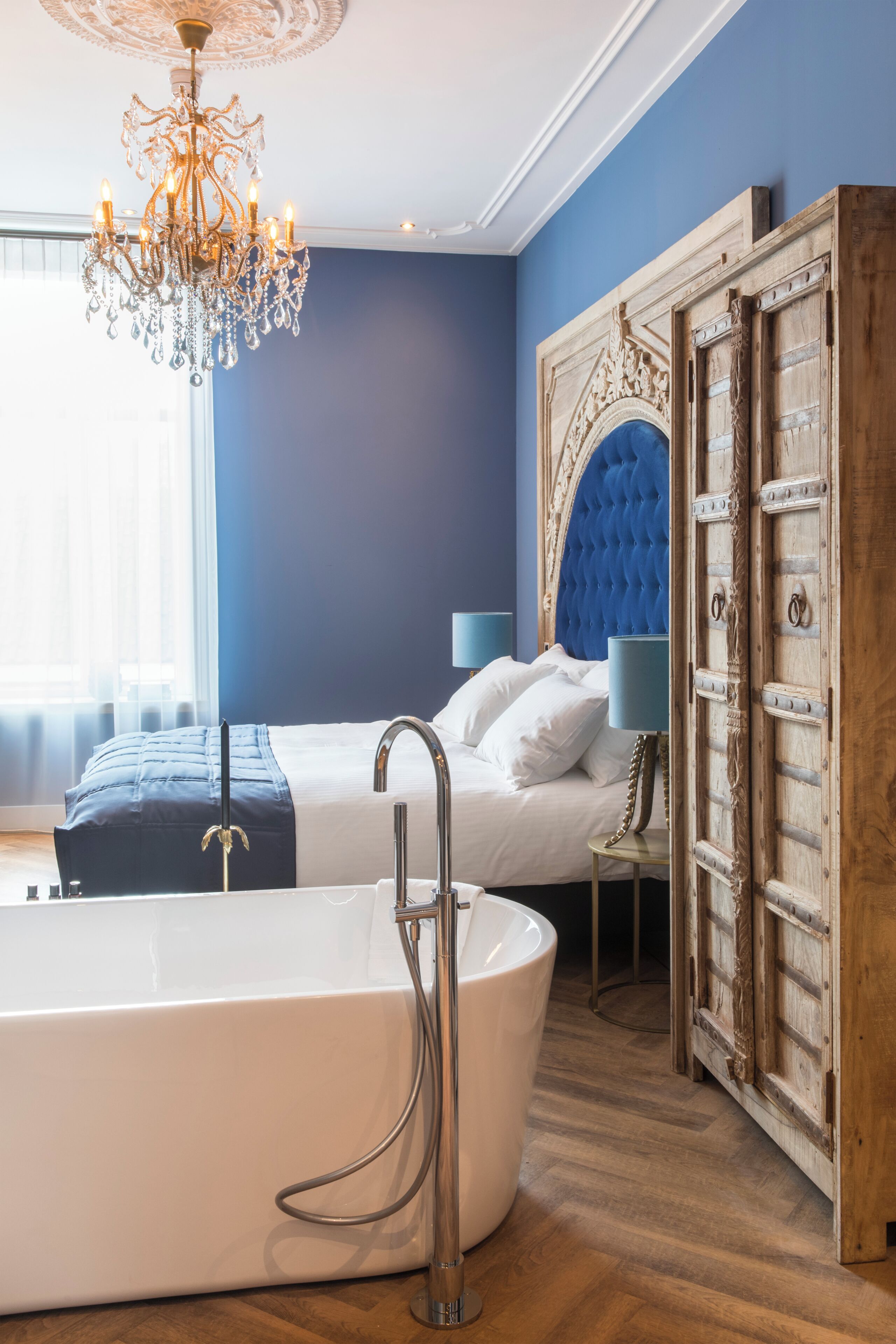 Un dormitorio con una bañera grande, una cama con cabecera capitonada y un candelabro.
