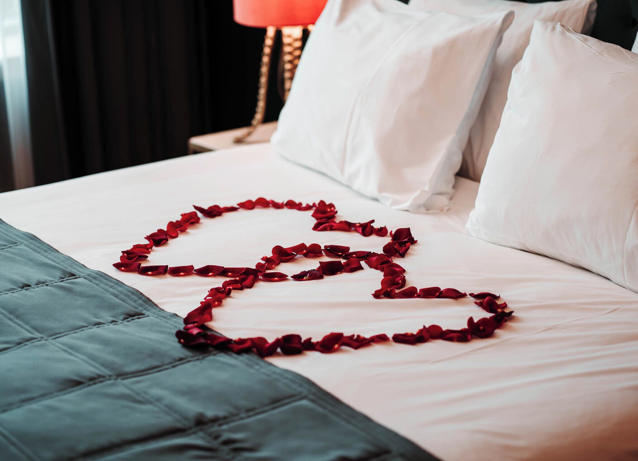 A bed with white bedding and a heart-shaped arrangement of red rose petals.