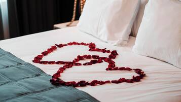 Una cama con ropa blanca y un arreglo de pétalos de rosa roja en forma de corazón.