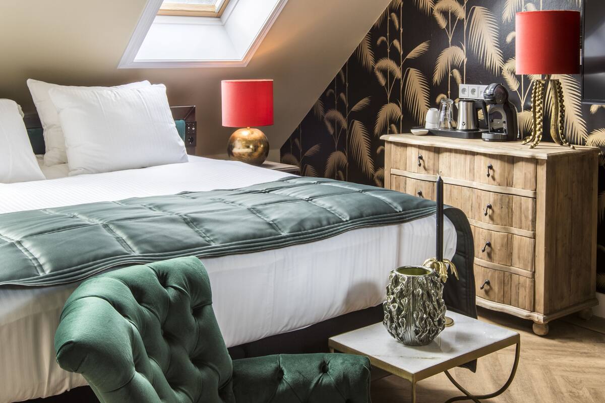 A bedroom with a bed, a green armchair, a wooden dresser, and a red lamp.