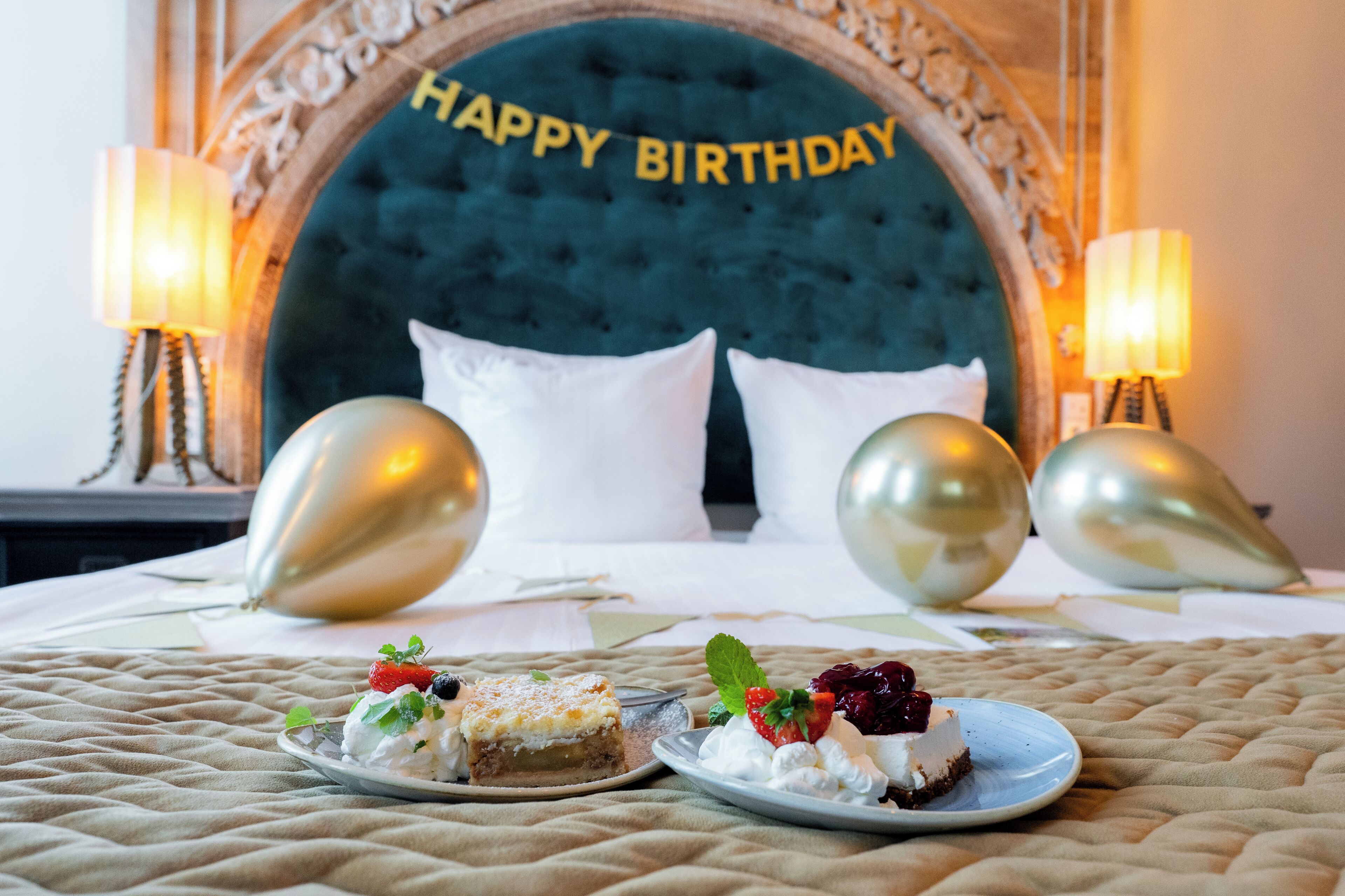 Una cama con un cabecero que dice 'Feliz Cumpleaños', dos mesitas de noche con lámparas y dos platos de postre sobre la cama.
