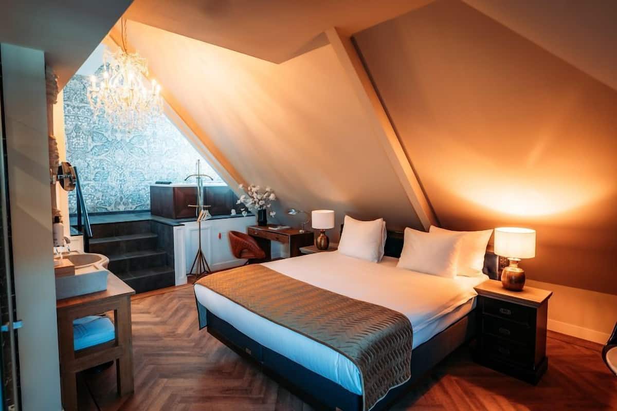 A bedroom with a large bed, a desk, a chair, a nightstand, and a chandelier.