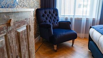 Un dormitorio con un sillón tapizado oscuro, suelo de madera, cama con ropa de cama blanca y una ventana con cortinas traslúcidas.