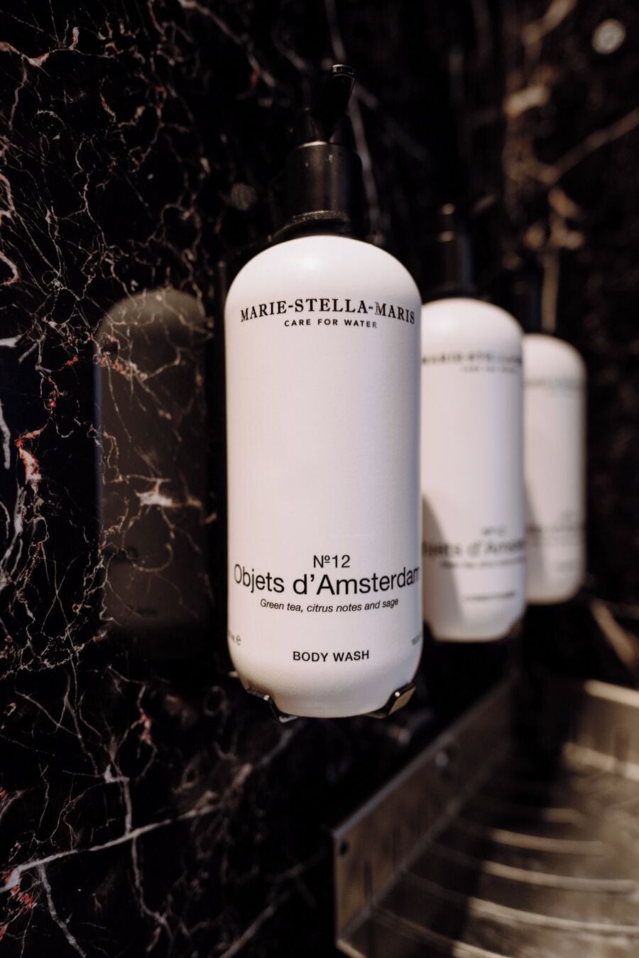 Une série de flacons blancs, tous étiquetés, dont l’un affiche en évidence les mentions « Marie-Stella-Maris, soins pour l’eau » et « Objets d’Amsterdam, gel douche au thé vert, aux notes d’agrumes et à la sauge ».