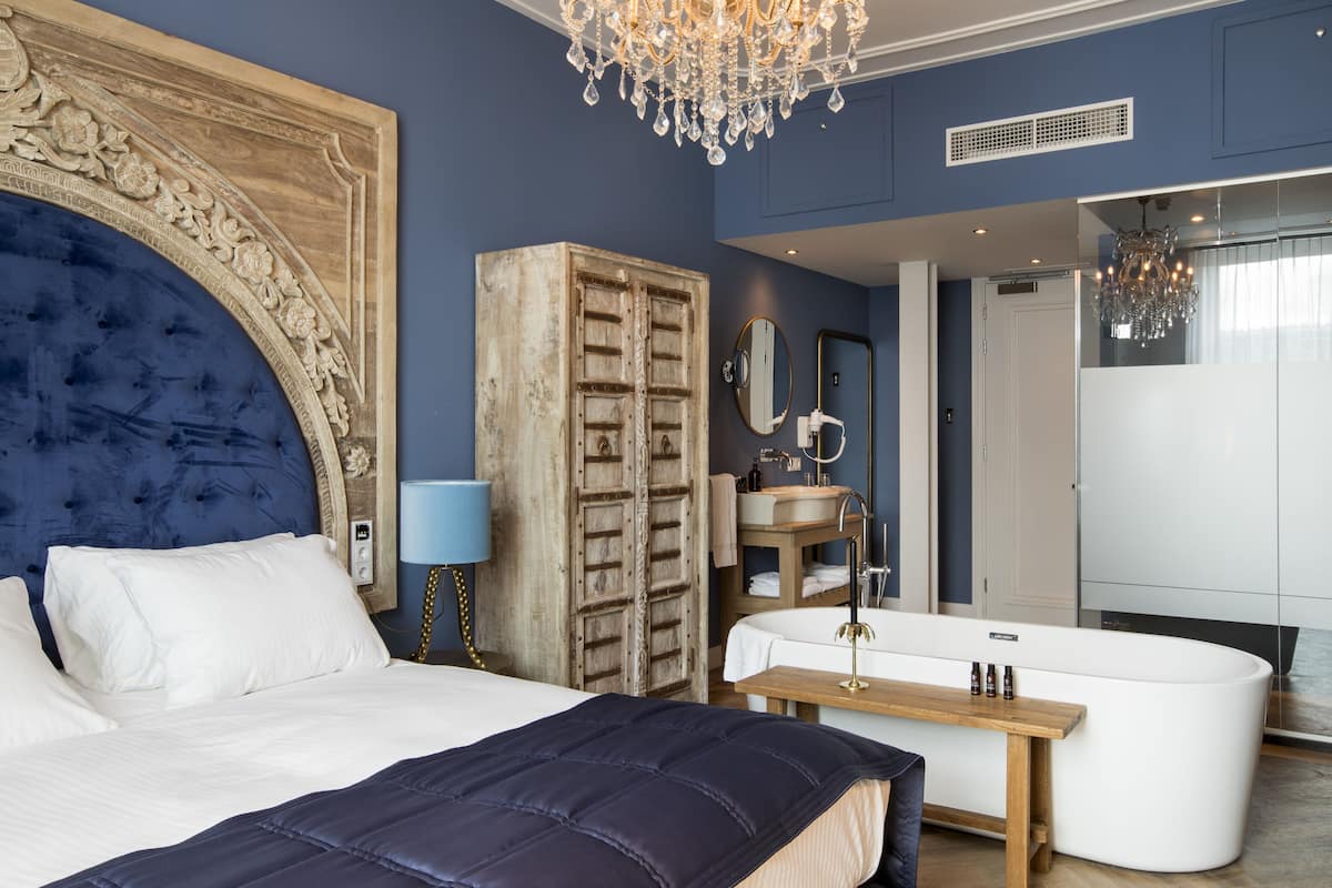 A bedroom with a large bed, a wooden wardrobe, a bathroom with a bathtub, and a chandelier.