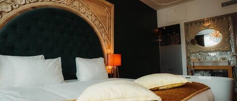 Una cama grande con cabecera oscura, almohadas blancas y una manta marrón. Arriba, un candelabro y en la pared, un espejo con marco ornamentado.